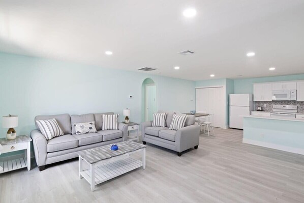 A modern living room with two sofas, a coffee table, and a kitchen area with white cabinets and appliances.