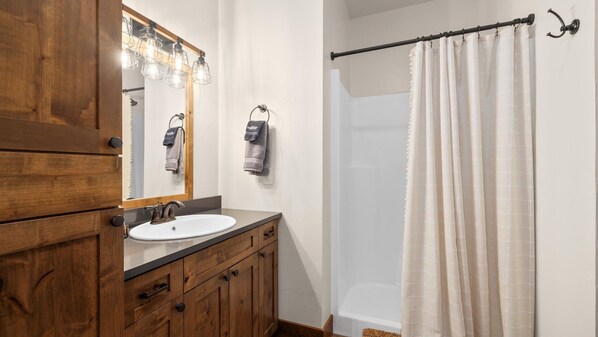 Une salle de bain avec un meuble-lavabo en bois, un lavabo blanc, un miroir avec éclairage intégré et une douche dotée d’un rideau beige.