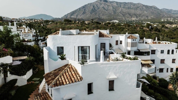A white building with a tiled roof, surrounded by greenery and a mountain in the background. - Lomas del Marbella | Puente Romano Penthouse (Marbella)