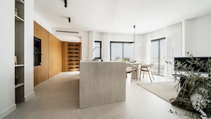 A modern kitchen with a central island, wooden cabinets, and a dining area with a TV and plants. - Lomas del Marbella | Puente Romano Penthouse (Marbella)