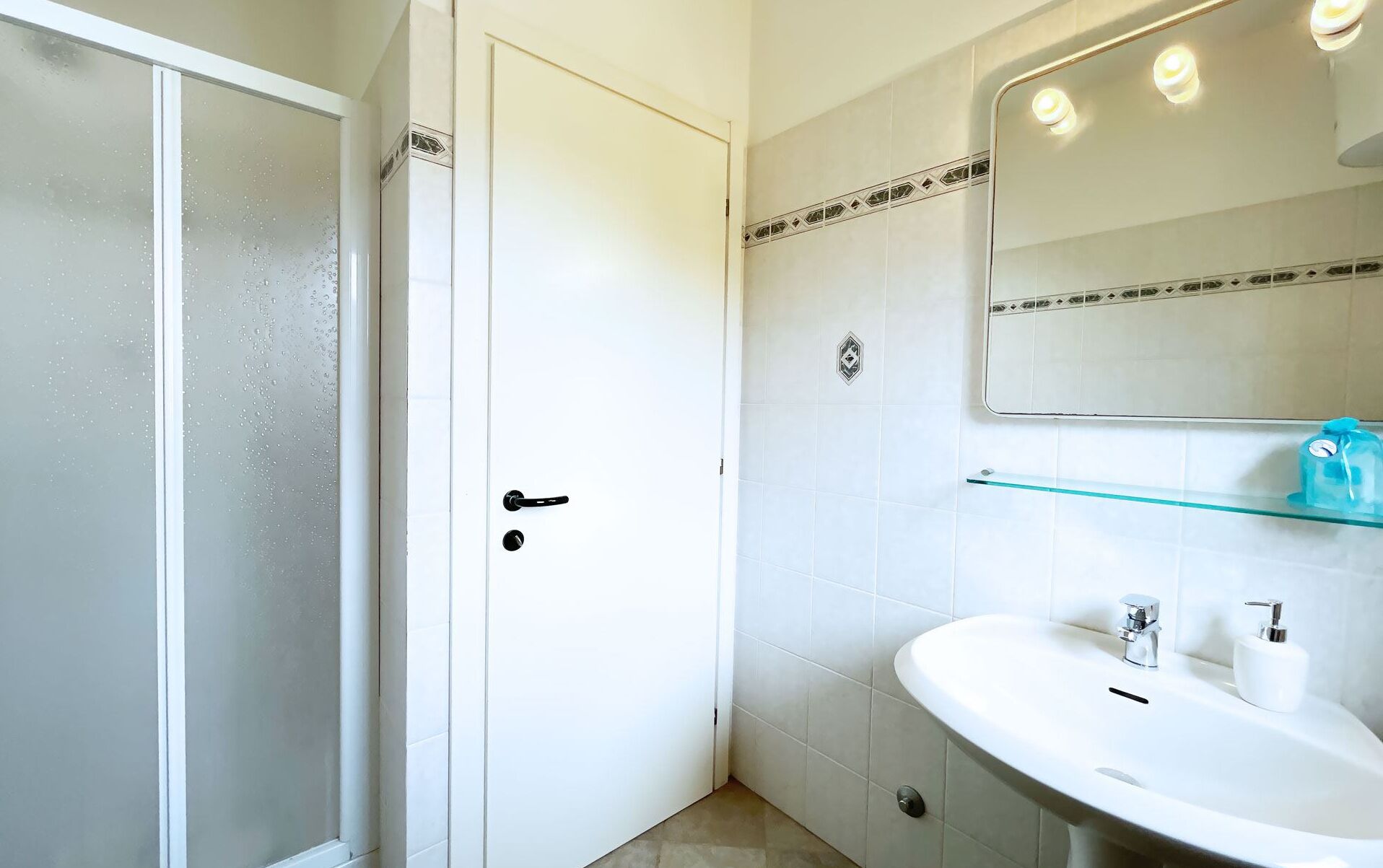 A bathroom with a white door, a glass shower enclosure, a white sink with a mirror above it, and a small shelf with toiletries.