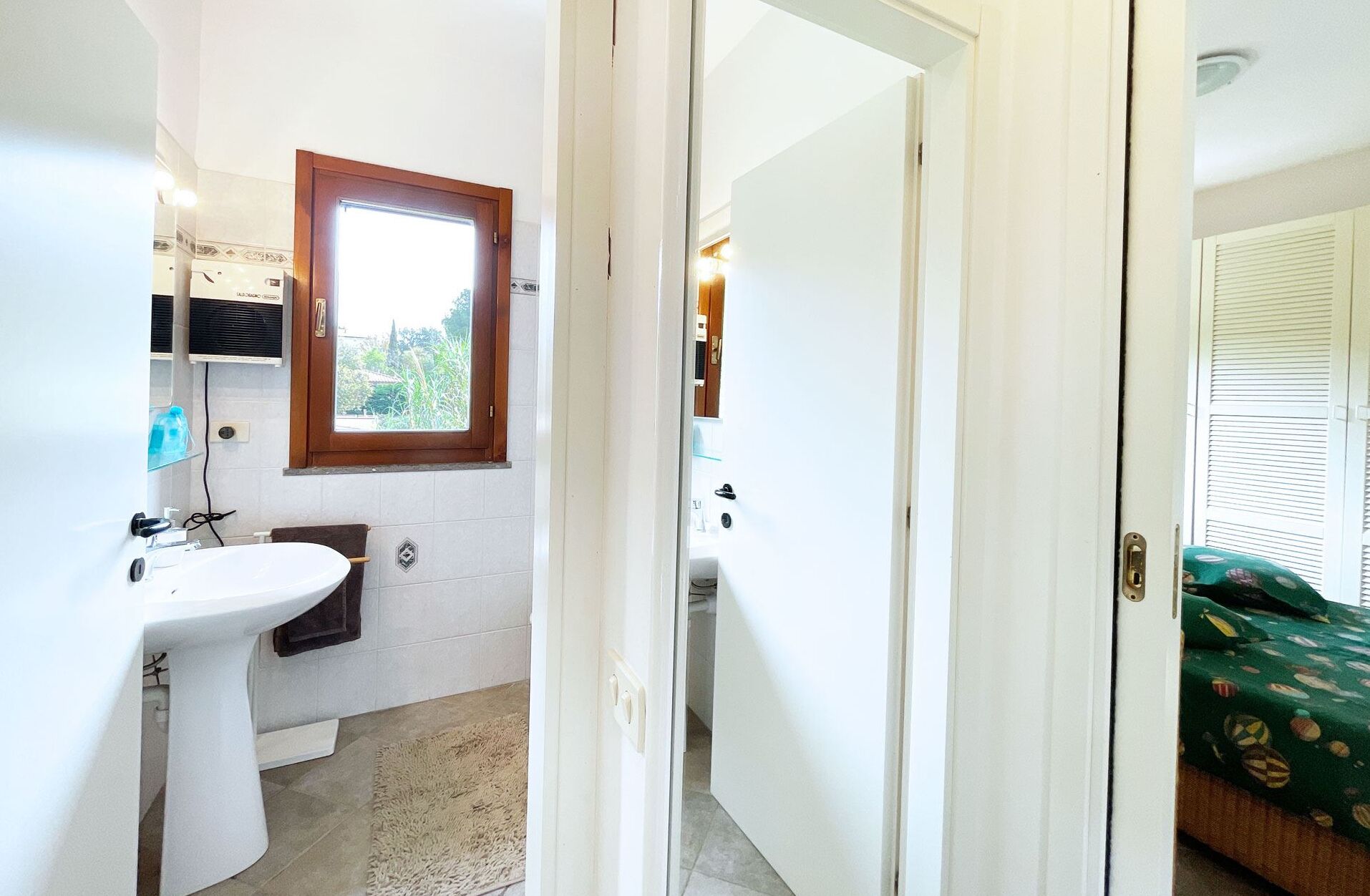 Un baño con un lavamanos blanco, una ventana y una puerta que da a una habitación con una cama.