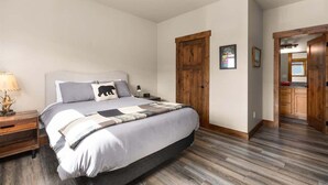 Een slaapkamer met een groot bed, houten vloer, nachtkastje, lamp en een deur die naar een keuken leidt.