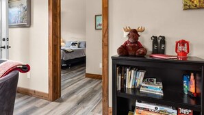 Een kamer met een houten vloer, een boekenplank met boeken en een rendierbeeldje, een bed met een grijze deken, en een schilderij aan de muur.