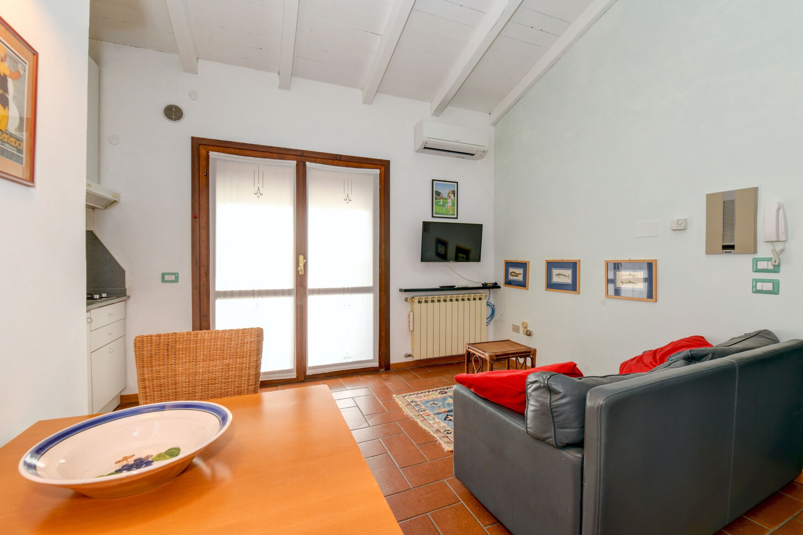 Una sala de estar con sofá, una mesa de comedor con un bol, una silla de mimbre, un ventanal corredero, un televisor y un radiador.