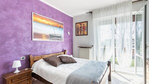 Et soveværelse med en seng, et natbord, en lampe, et vindue med gardiner og et billede på væggen.