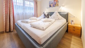 A neatly made bed with a grey headboard, a bedside table with a book, and a window with sheer curtains. - Ferienwohnung Unternberg 45qm, Terr., Küche, Behinderteng., 1 Schlafzimmer (Ruhpolding)