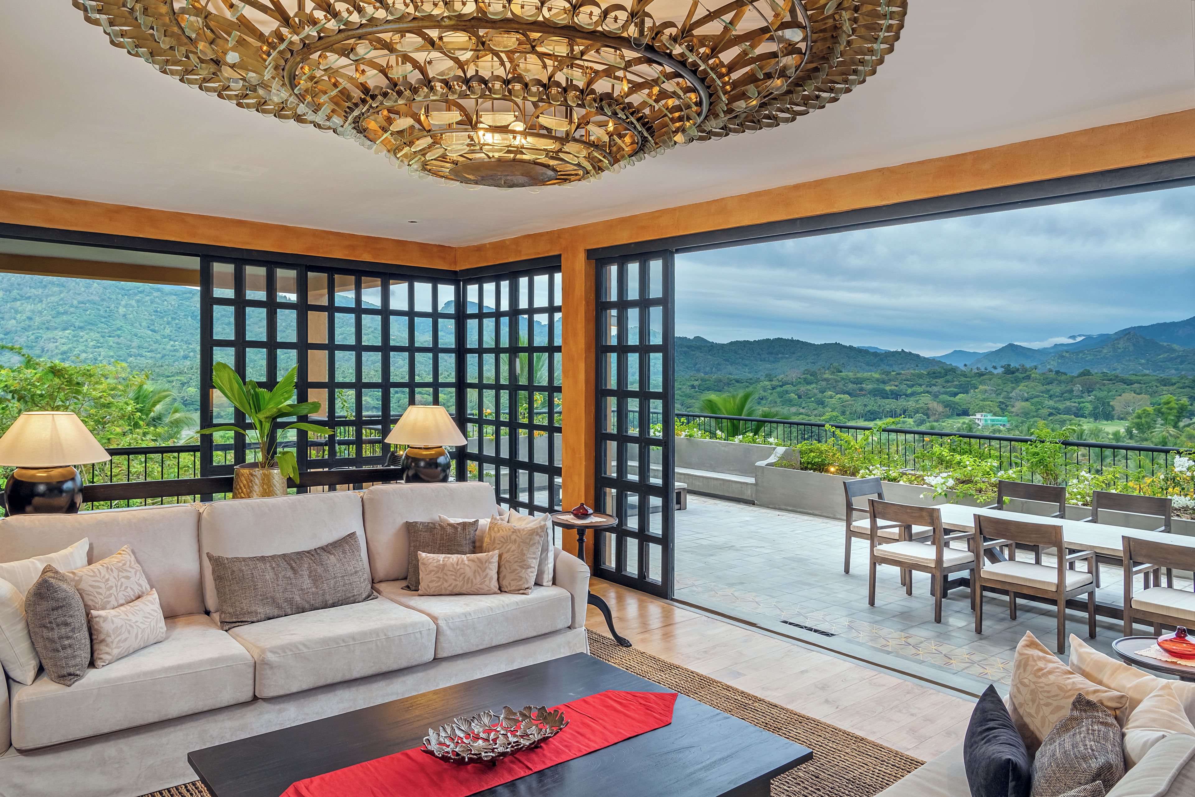 A modern living room with a sofa, coffee table, and dining area, featuring a large window with a mountain view.