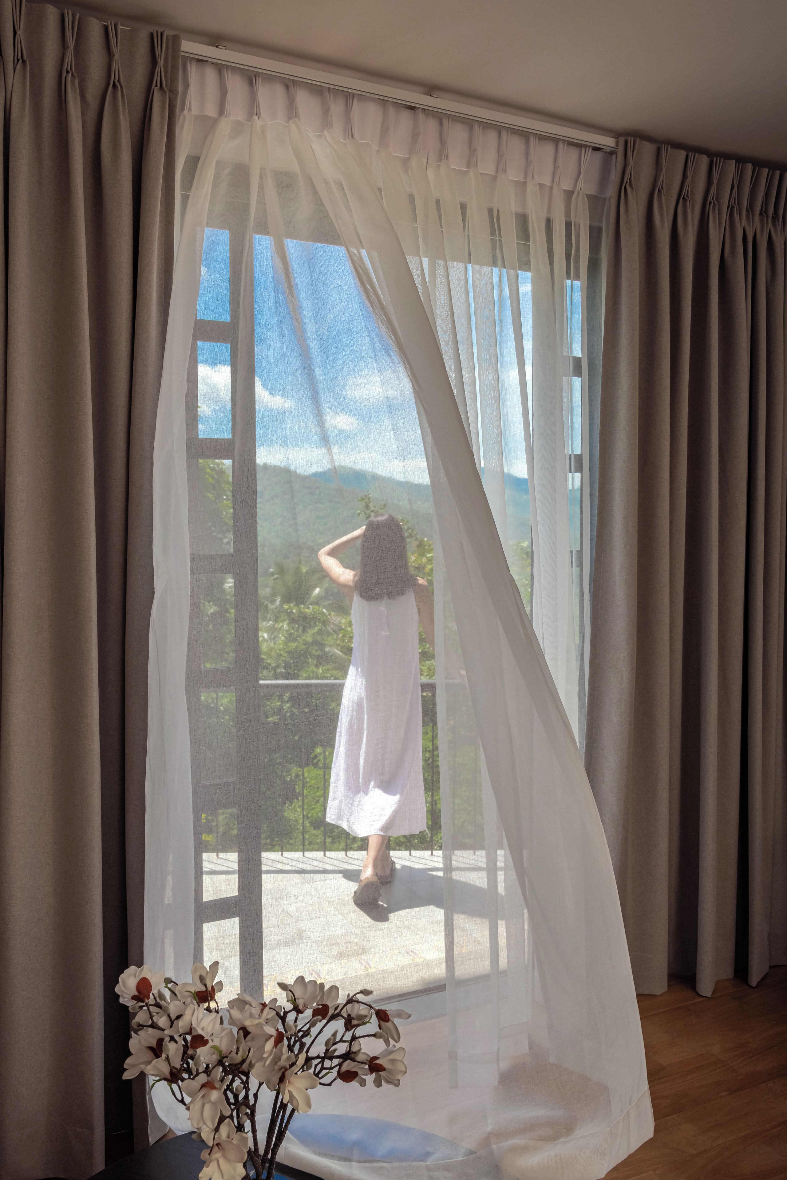 Ett rum med ett stort fönster som är täckt av tunna, genomskinliga gardiner, en krukväxt och utsikt över grönområden utanför.