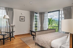 Habitación de hotel con cama, escritorio, silla, ventana con vista a follaje y un cuadro en la pared.