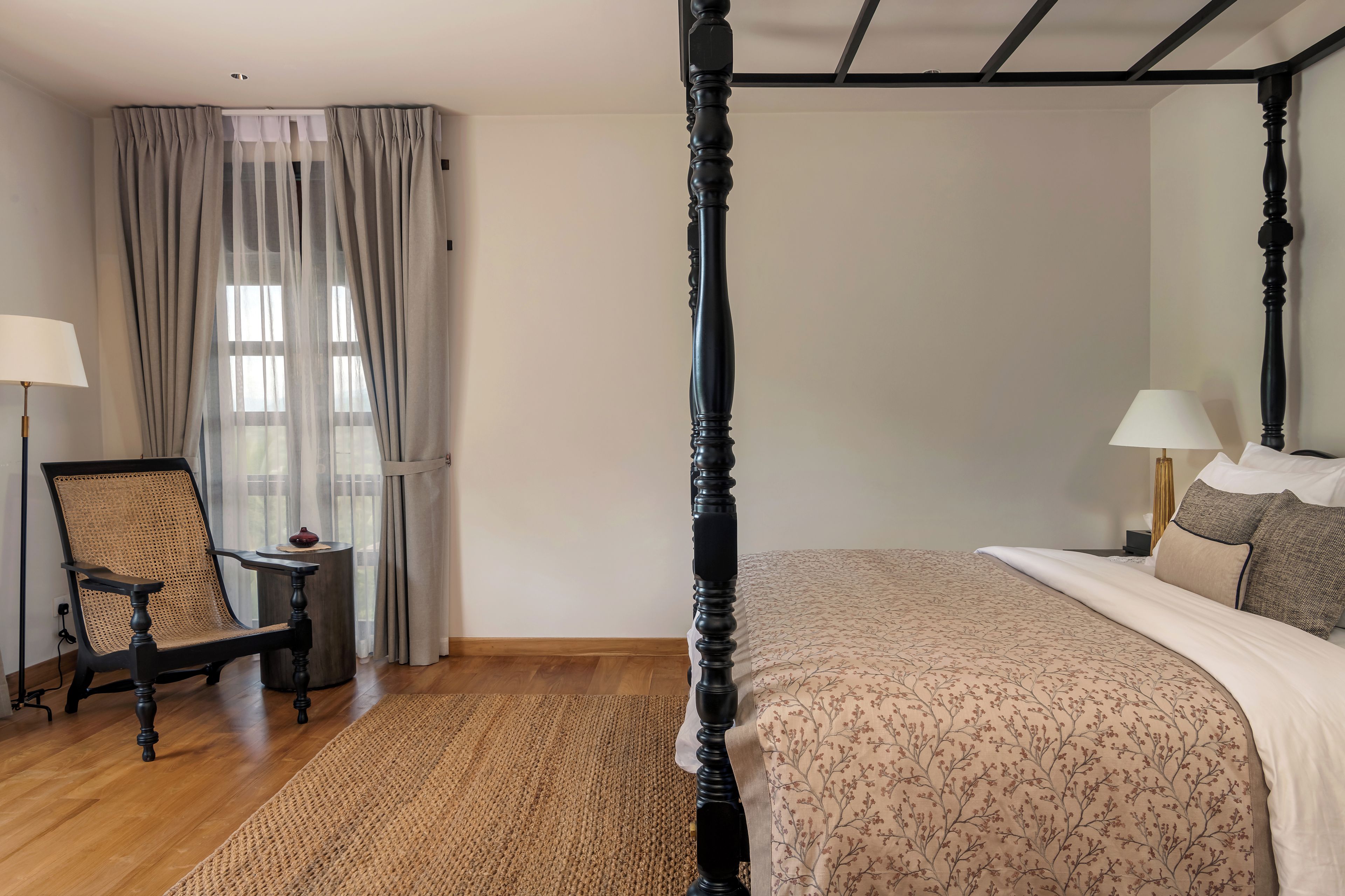 A bedroom with a four-poster bed, a chair, a small table, and a window with curtains.