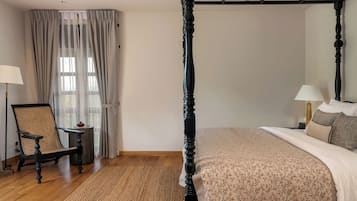 A bedroom with a four-poster bed, a chair, a small table, and a window with curtains.