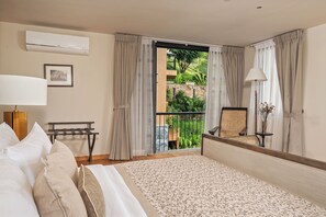 Habitación de hotel con cama, escritorio, silla, lámpara y vistas a un jardín desde un balcón.