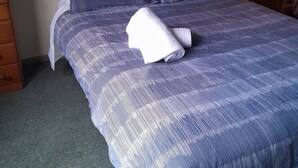 Un letto rifatto con cura, coperto da una trapunta a motivi blu e bianchi, due cuscini e un asciugamano bianco arrotolato. Accanto al letto, un comodino in legno con una lampada accesa.