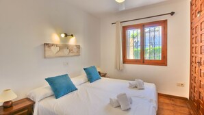 A neatly made bed with blue pillows, a wooden headboard, and a window with curtains. - Bungalow in Denia 3 minutes from the beach with air-conditioning (Denia)