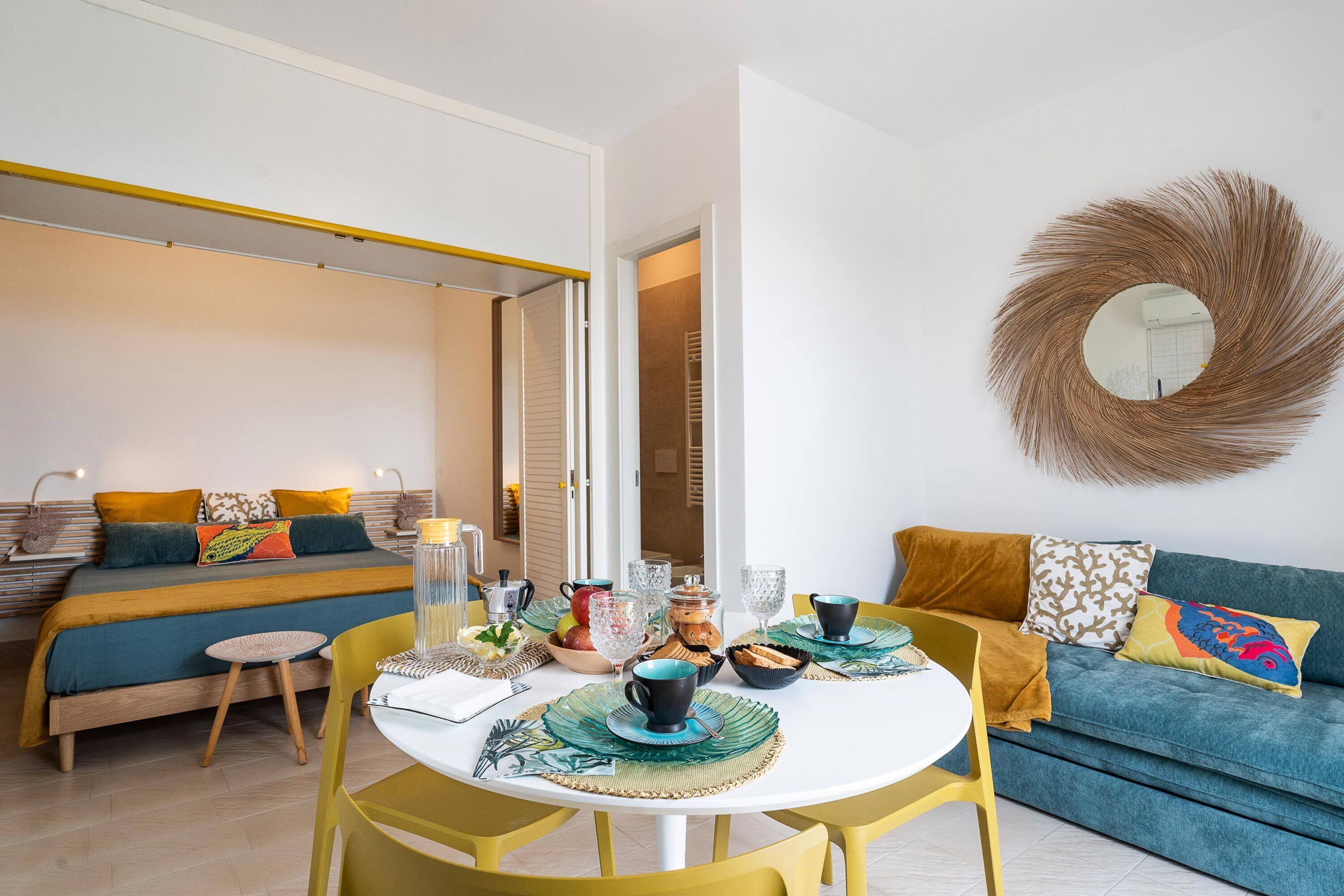Een moderne woonkamer met een bank, een eettafel met stoelen en een bed met kleurrijk beddengoed.
