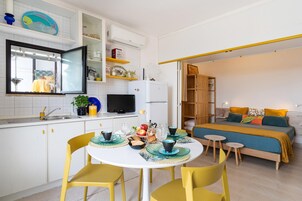 Una cucina compatta con mobili bianchi, un tavolo da pranzo con sedie gialle, e un letto con copriletto blu e arancione nella stanza comunicante.
