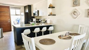 Een moderne keuken met witte kasten, een centraal eiland met zitplaatsen, en een eethoek met een witte tafel en stoelen.