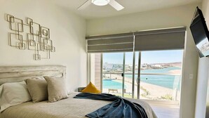 Een slaapkamer met een bed, een plafondventilator, een groot raam met uitzicht op een strand en zwembad, en een aan de muur bevestigde televisie.