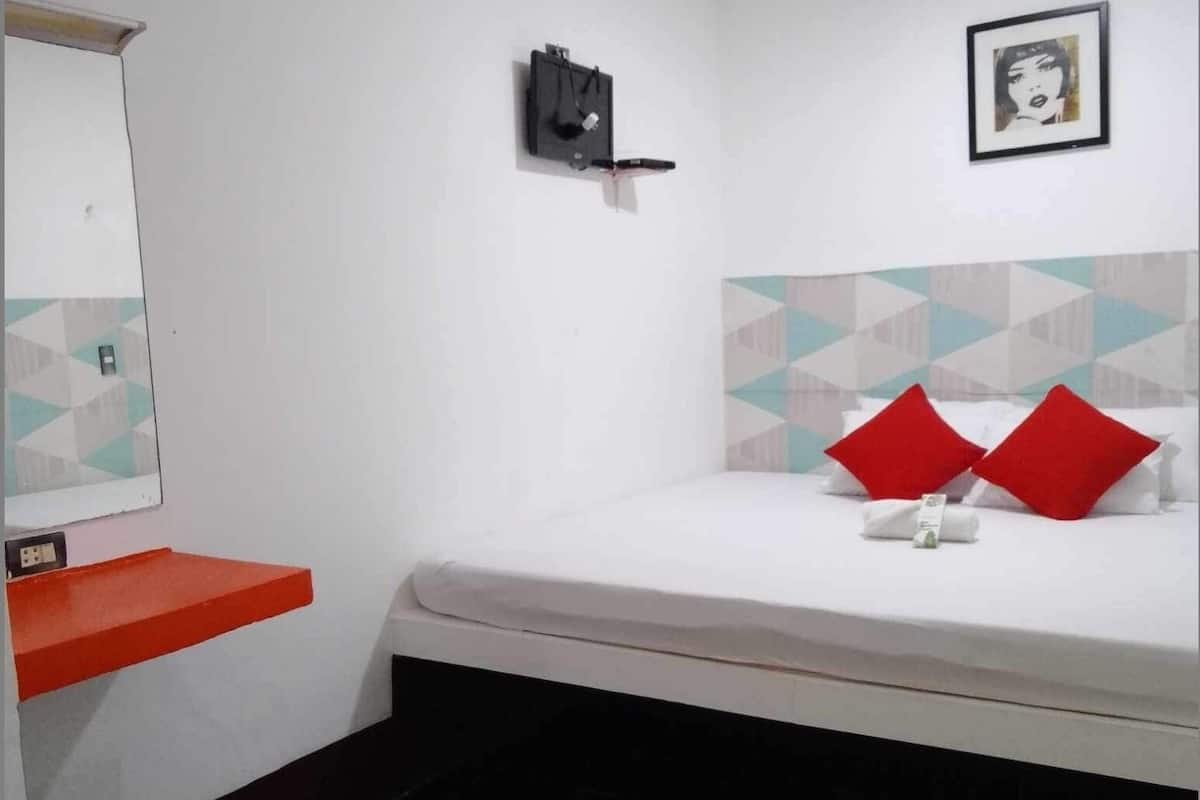 A small, clean room with a bed, a red shelf, a wall-mounted light, and a framed picture.