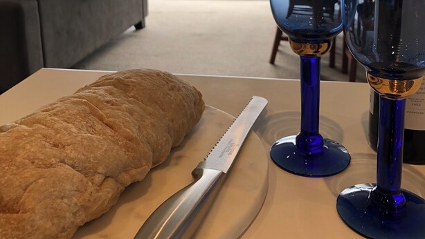 A table with a loaf of bread, a knife, two wine glasses, and a bottle of wine in a room with a view of a vineyard and mountains through large windows. - Ben Morven Vineyard Cottage (Fairhall)
