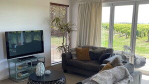 A modern living room with a grey sofa, a coffee table, a TV on a glass stand, and a large window with a vineyard view. - Ben Morven Vineyard Cottage (Fairhall)