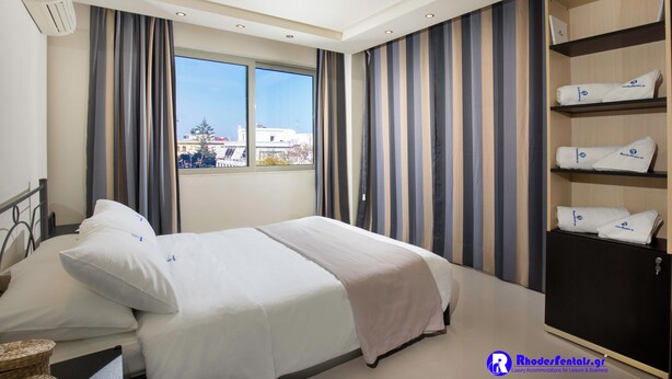 A hotel room with a bed, a wardrobe, a window, and a view of buildings outside. - Rhodes Central Suite (Rhodes)