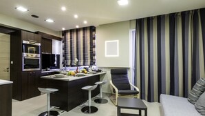 A modern kitchen with built-in appliances, a dining area with bar stools, and a living space with a sofa and striped curtains. - Rhodes Central Suite (Rhodes)