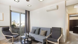 A modern living room with a grey sofa, two armchairs, a coffee table, and a view of the city through large windows. - Rhodes Central Suite (Rhodes)