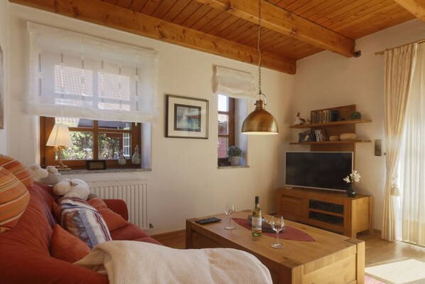 Uma sala de estar acolhedora com teto de madeira, um sofá com almofadas listradas, uma mesa de centro de madeira com uma garrafa de vinho e copos, uma televisão sobre um suporte de madeira e uma janela com persiana branca.