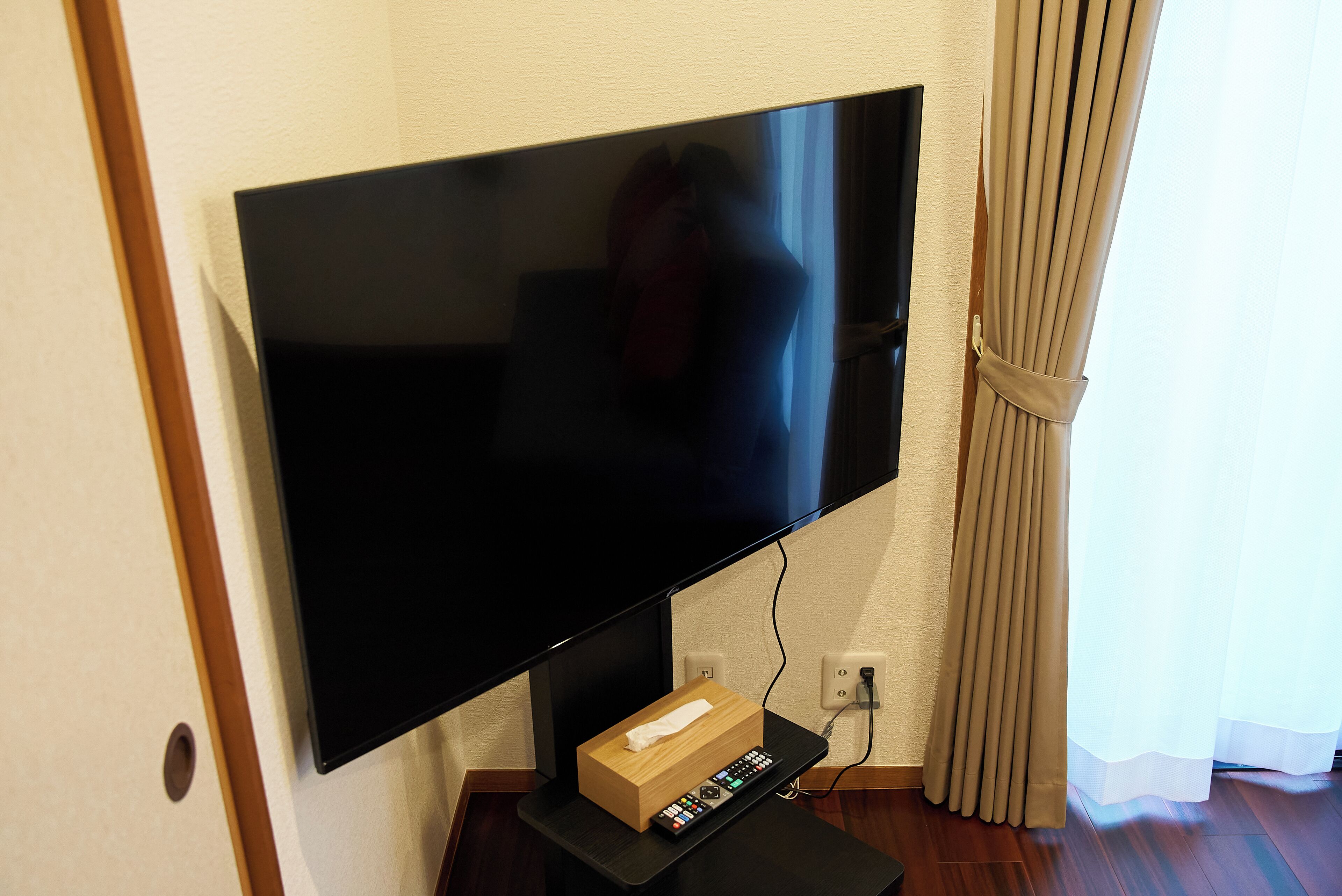 A flat-screen TV mounted on a wall, a remote control box on a stand below it, and a curtain partially drawn to the side.
