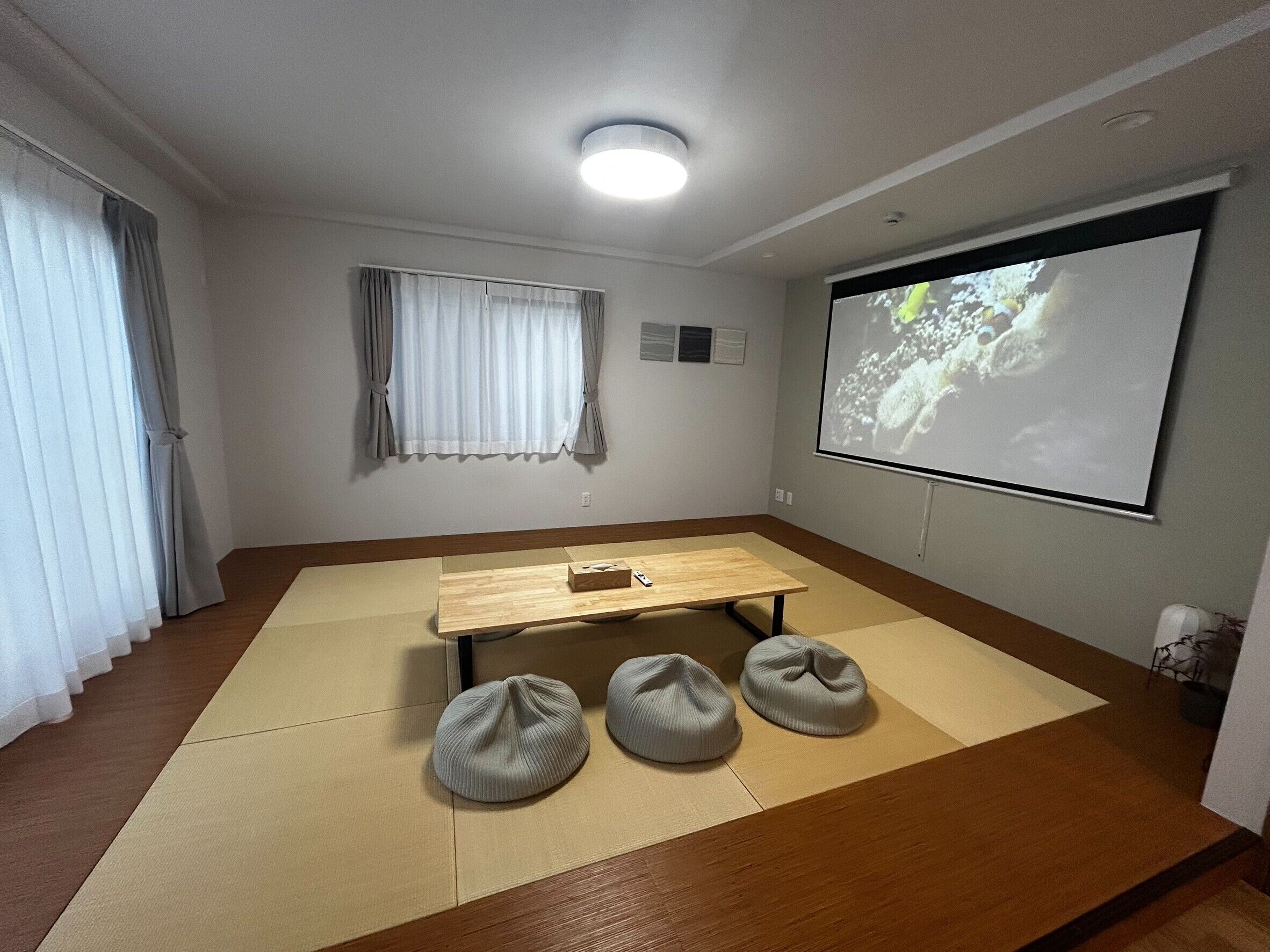 A room with a wooden table, bean bag chairs, a projector screen, and a window with curtains.