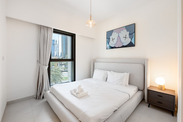 A modern bedroom with a large bed, a bedside table with a lamp, a window with curtains, and a wall-mounted artwork.