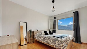 A bedroom with a large bed, a wooden dresser, a mirror, and a window with a mountain view.