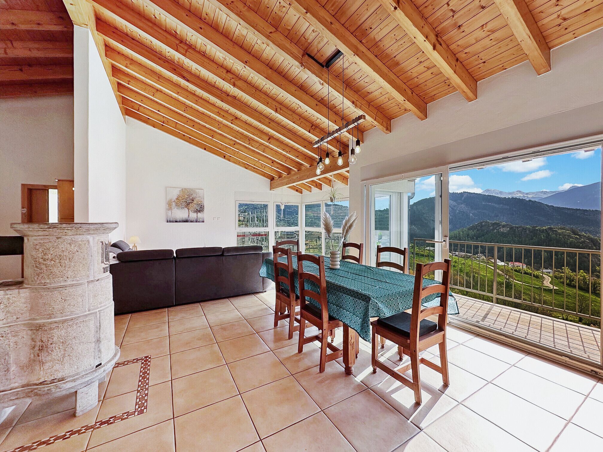 Un espace repas comprenant une table et des chaises, un pilier en pierre, un canapé et une vue sur les montagnes, visible à travers de grandes fenêtres.