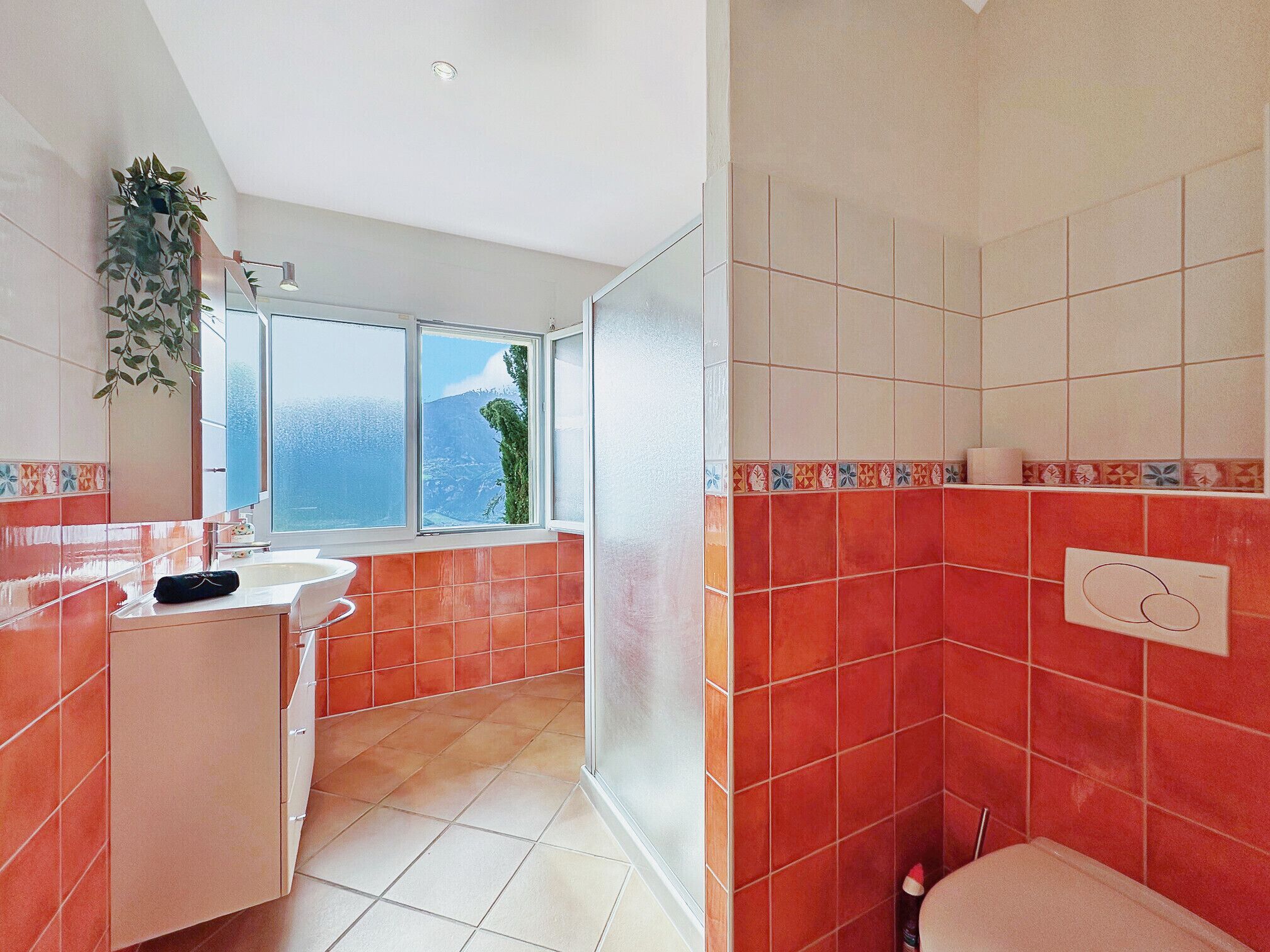 Un baño con baldosas rojas, un lavamanos blanco y una ventana con vistas a montañas.