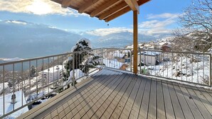 A wooden deck with a metal railing overlooking a snowy landscape with houses and mountains in the background.