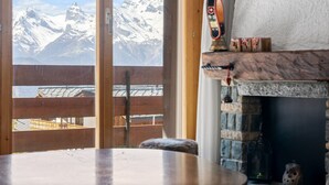 A room with a view of snow-capped mountains through a large window, a wooden table, and a stone fireplace with a decorative bell. - Le Torrent 16 - 6 pers - au calme (Haute-Nendaz)