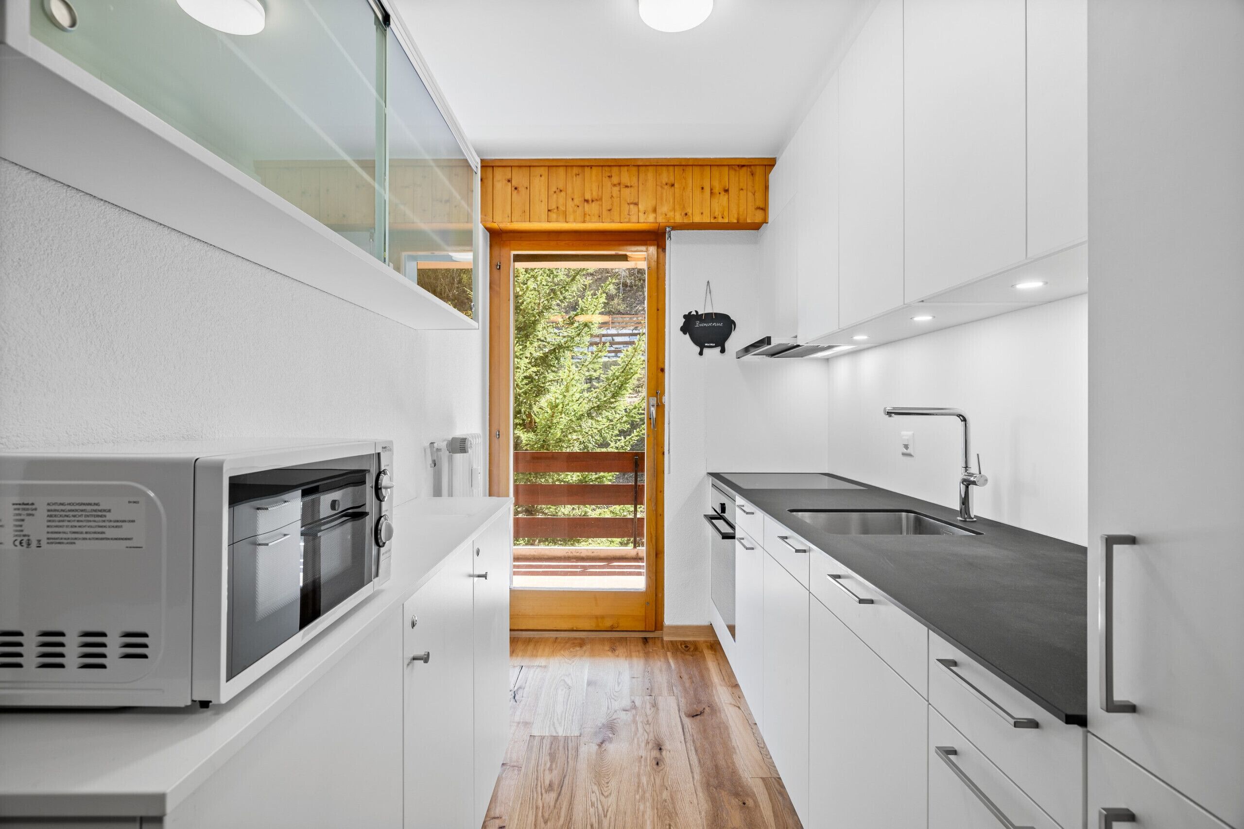 A modern kitchen with white cabinets, a built-in oven, and a large window overlooking a wooden deck.