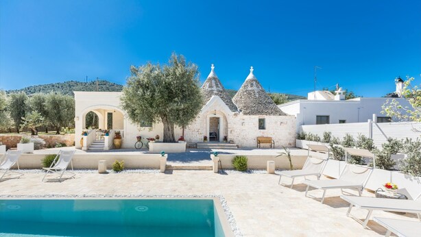 A poolside area with a stone pool, lounge chairs, and a building with a white facade and two conical roofs. - 3856 Trulli Ulivo di Puglia by Barbarhouse (Fasano)