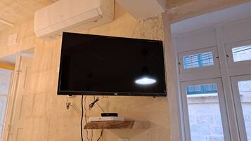 Ein flachbildfernseher an der Wand, darüber eine weiße Klimaanlage und darunter ein Holzregal mit einem Gerät.