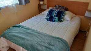 Ein Bett mit weißer Decke, grüner Wolldecke, einem gemusterten Kopfkissen und einem hölzernen Kopfteil.