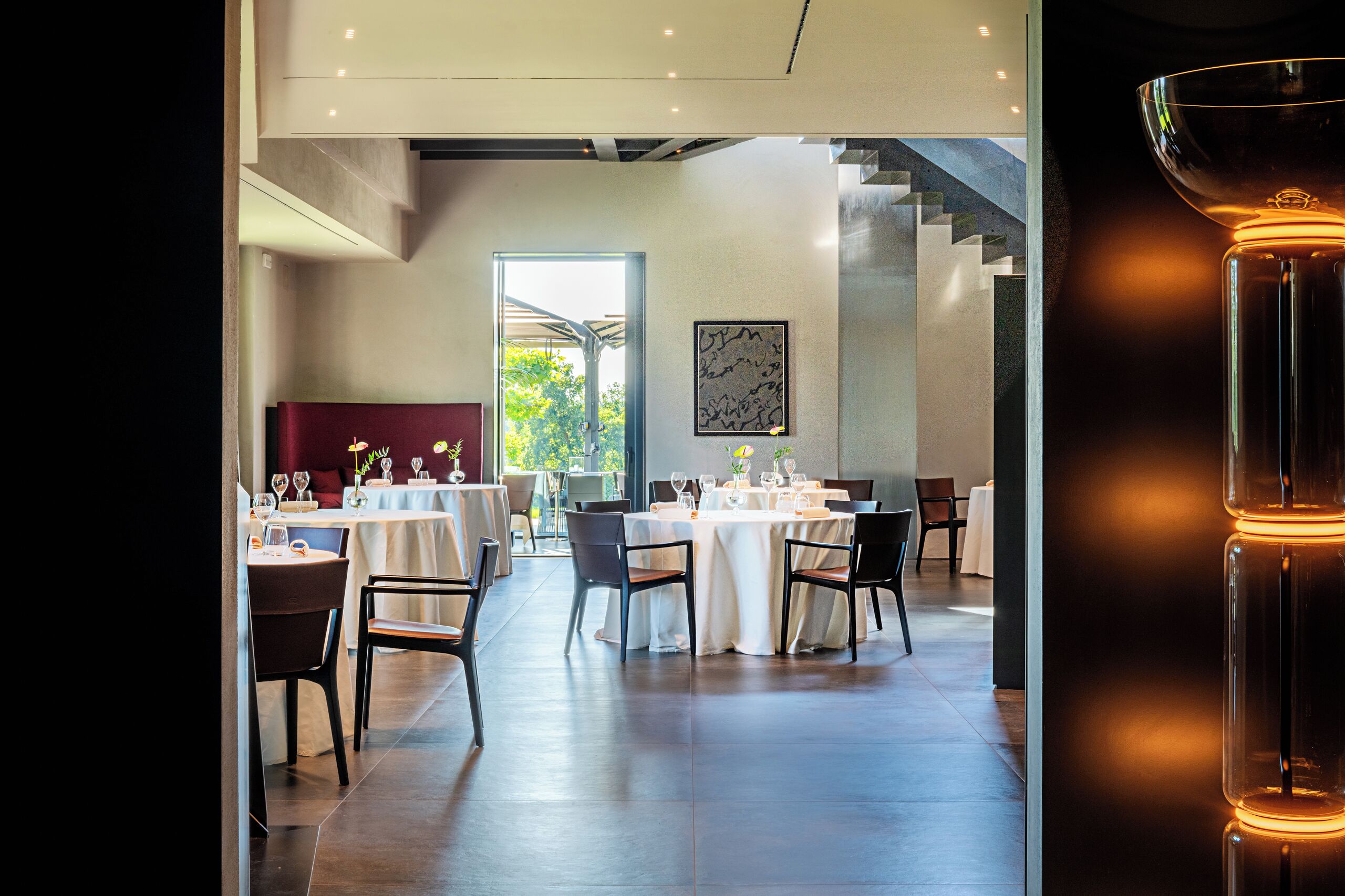 Um restaurante moderno com mesas preparadas para refeições, uma escadaria e vista para o verde lá fora.