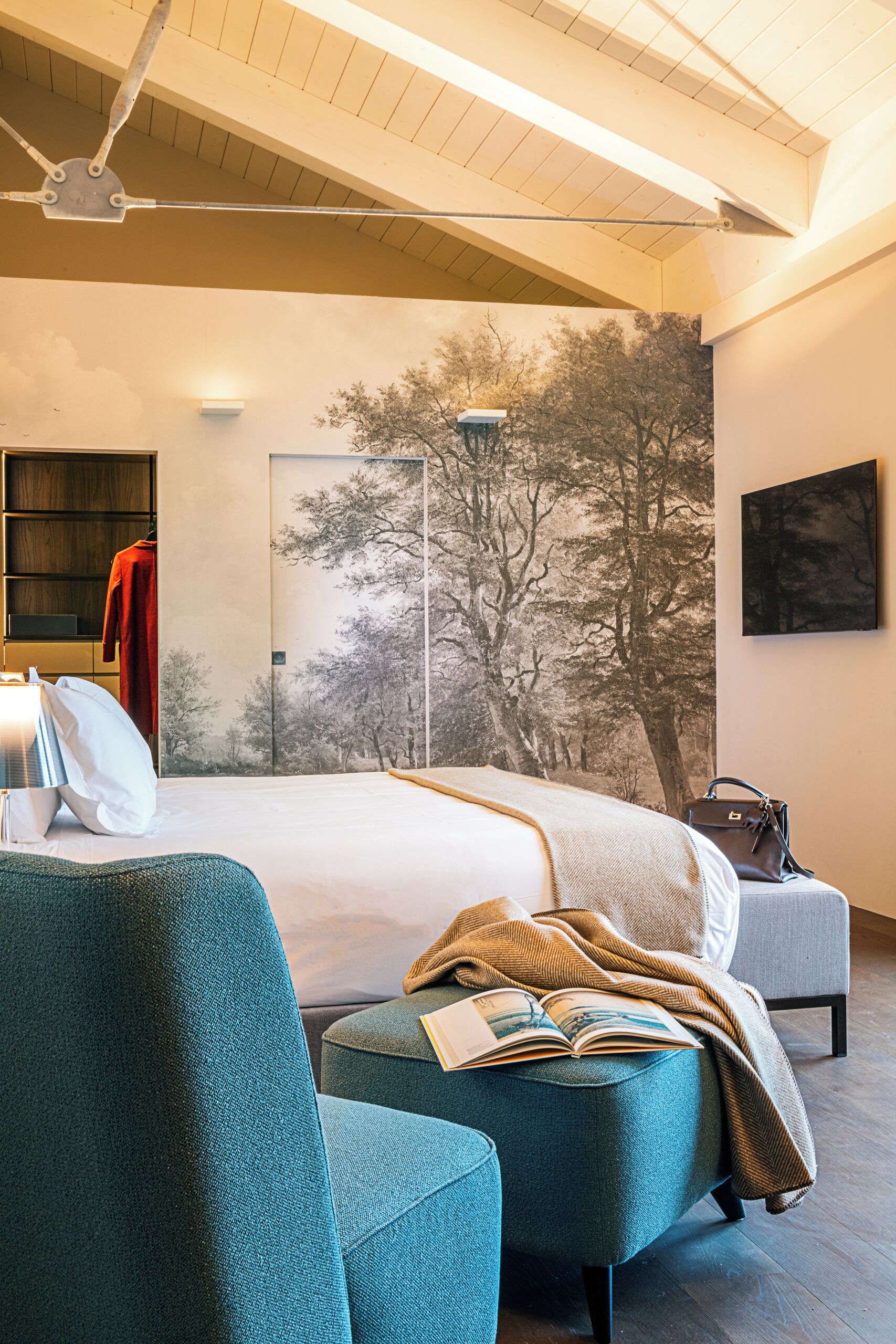 A modern hotel room with a large bed, a seating area with a book, a wardrobe, and a painting of trees.