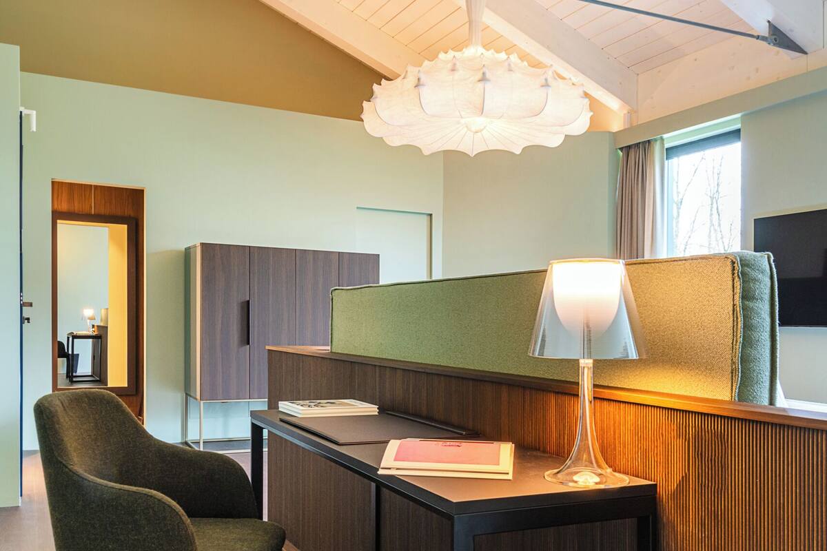 A modern hotel room with a wooden ceiling, a desk with a lamp, a chair, and a view of the outdoors.