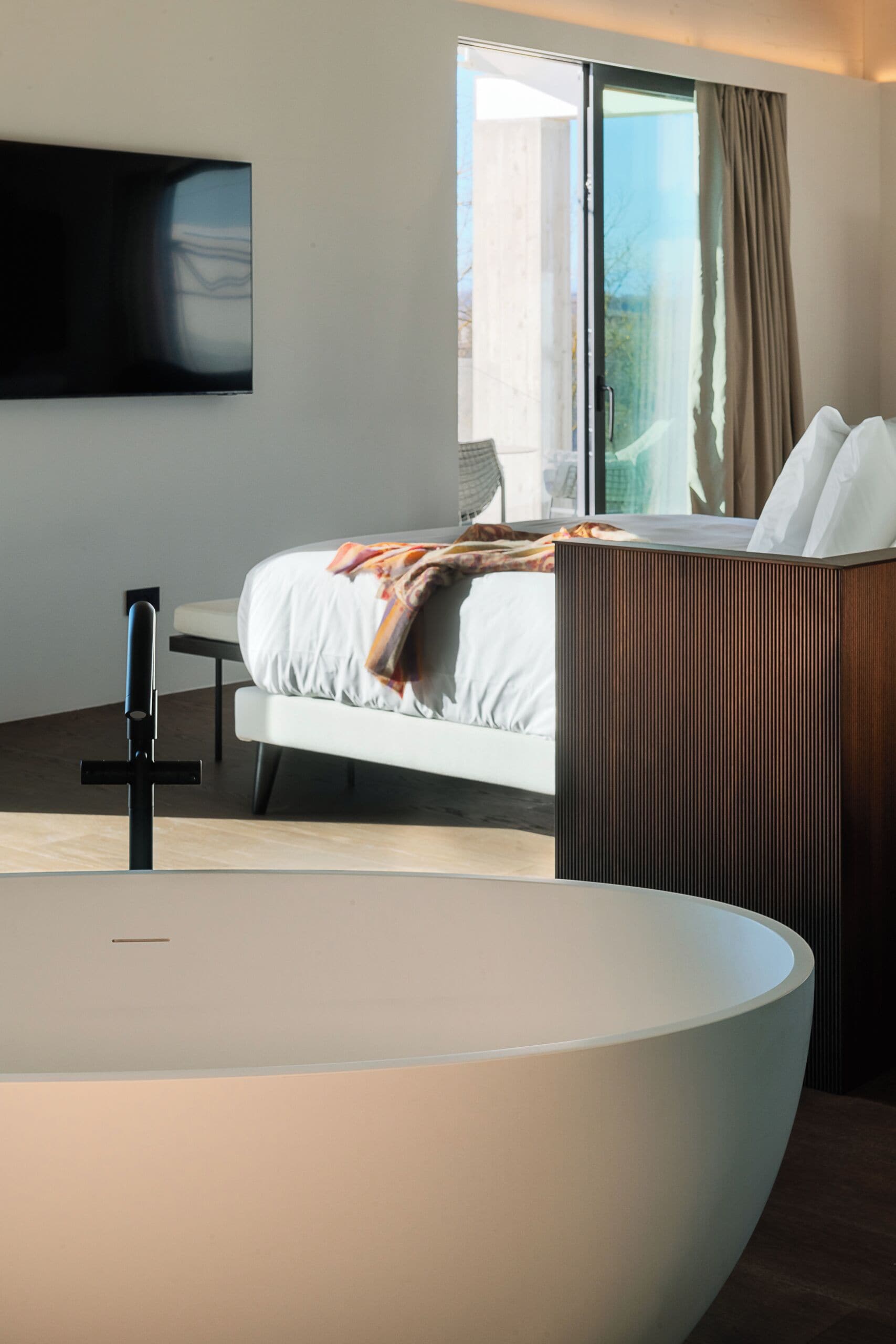Una habitación de hotel moderna con una cama grande, una mesita de noche, un televisor de pantalla plana en la pared y un amplio ventanal con cortinas.