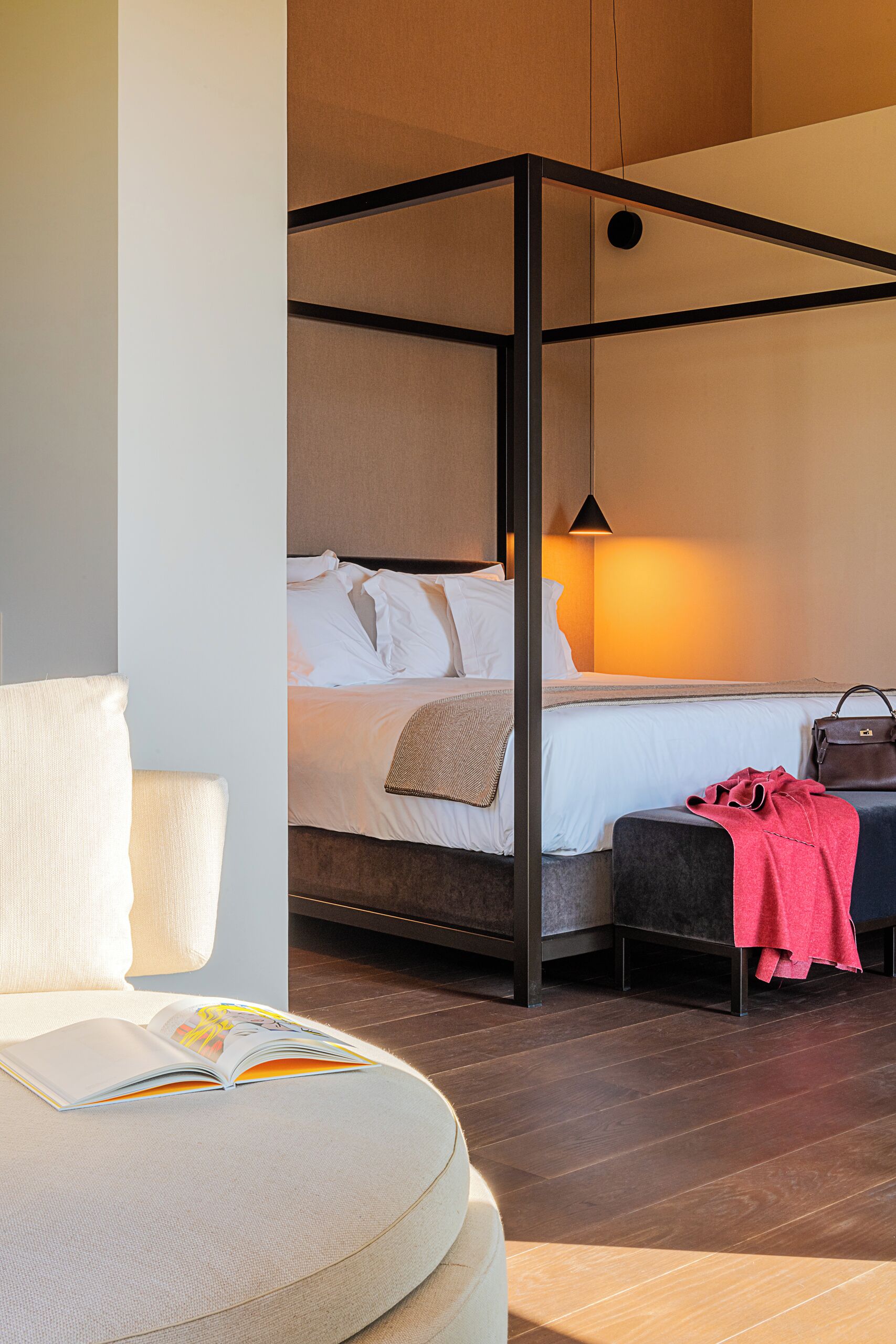 Ein modernes Hotelzimmer mit Himmelbett, Leseecke mit Sessel und Buch sowie einem kleinen Tisch mit einer Handtasche.