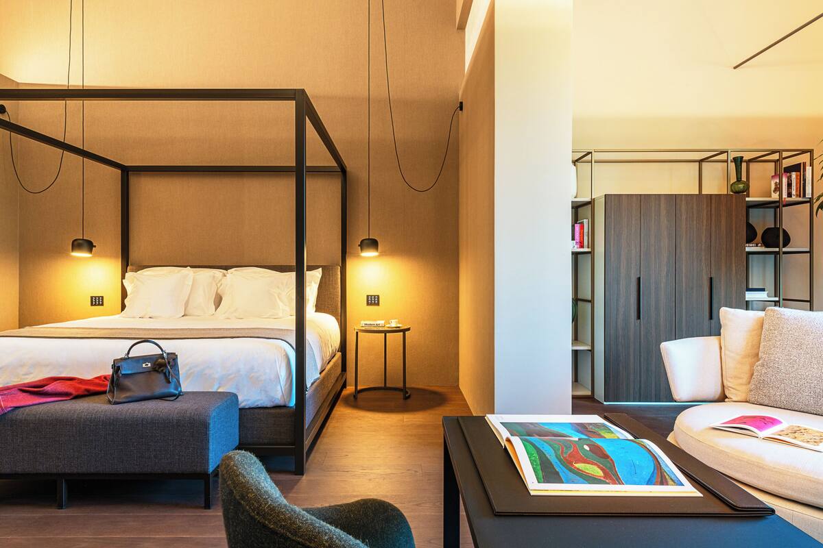 A modern hotel room with a four-poster bed, a bedside table, a reading area with a sofa and a coffee table, and a bookshelf.