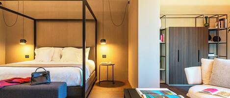 Una habitación de hotel moderna con una cama con dosel, una mesita de noche, una zona de lectura con un sofá y una mesa de centro, y una estantería.