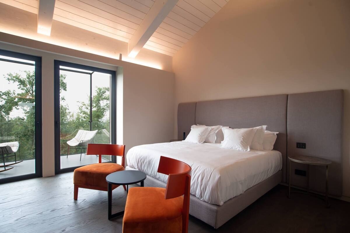 A modern bedroom with a large bed, two chairs, a small table, and a view of greenery through large windows.